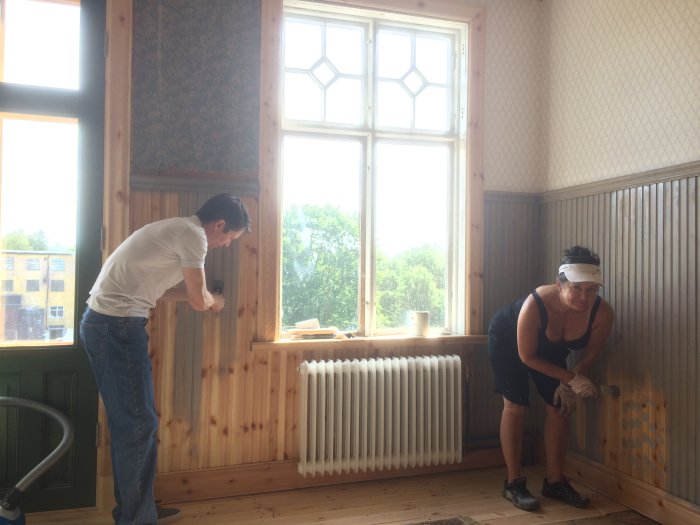 Två personer renoverar ett rum med träpaneler, stora fönster och en radiator.