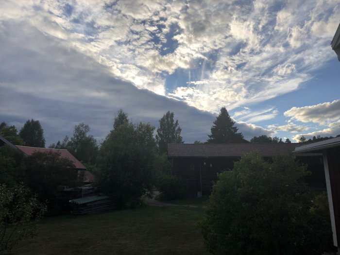 Molnfront över en trädgård med träd och hus, solstrålar bryter genom molnen.