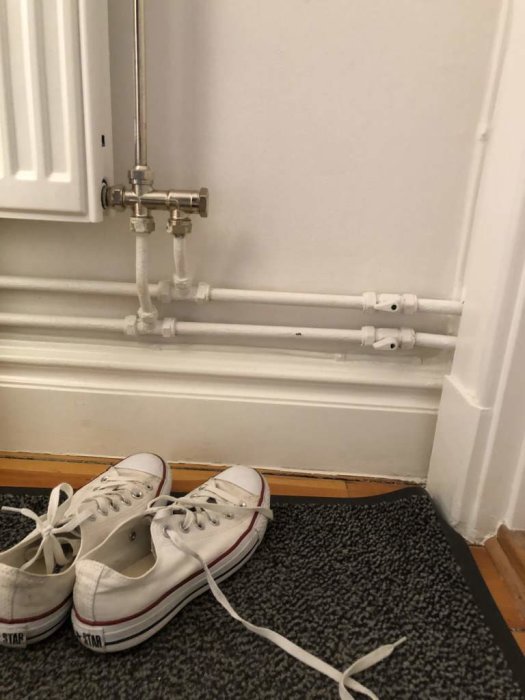 Värmeelement med termostatventil och anslutande rör vid en vit vägg, par vita sneakers framför.