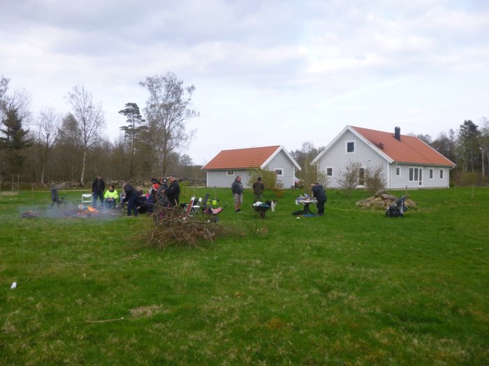 Människor samlade kring en säker eldplats i en trädgård med en villa i bakgrunden.