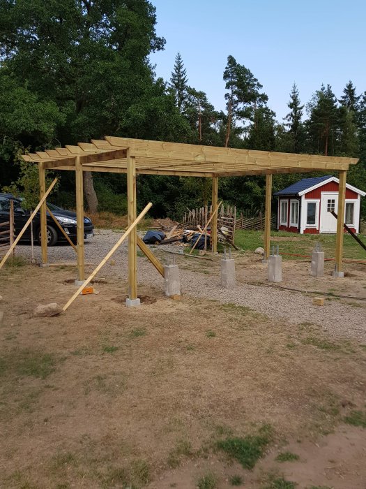 Nybyggd träpergola på betongfundament med stödstolpar och takbjälkar i en trädgård, omgiven av grönska och ett rött hus.