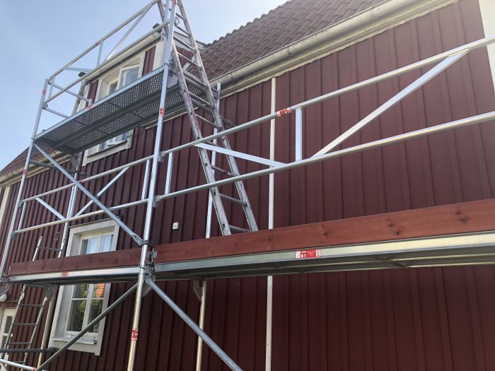 Byggställning monterad längs röd träfasad på ett hus under en klarblå himmel.