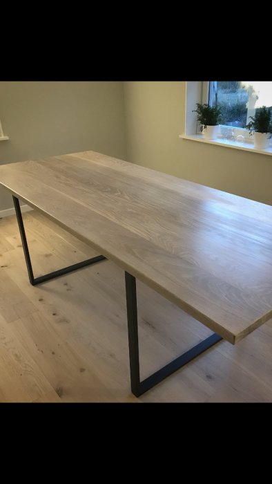 Ett enkelt träbord med modernt svart metallstativ på ett ljust trägolv mot en grå vägg.