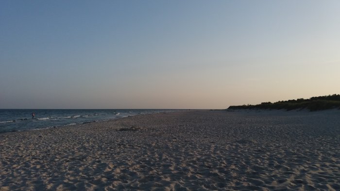 Solnedgång över en sandig strand vid havet med några personer och sanddyner i bakgrunden.
