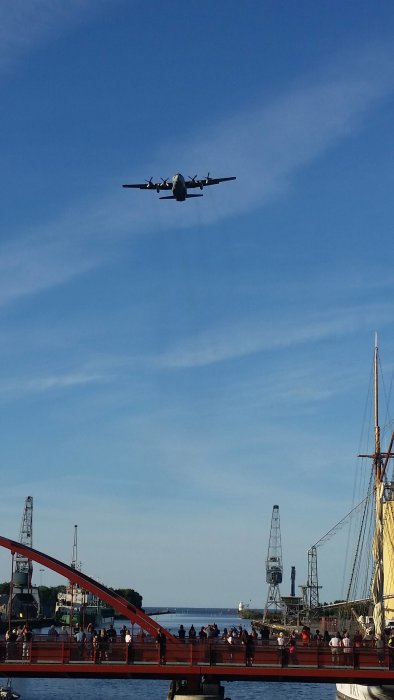 Hercules-flygplan flyger lågt över folkfyllt hamnområde i Lidköping vid solnedgång.