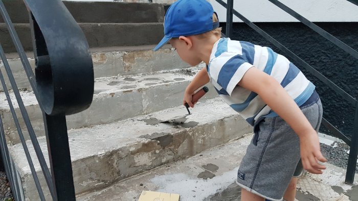 Fyraåring arbetar på betongtrappa med murslev, lappar håligheter, iklädd randig tröja och blå keps.