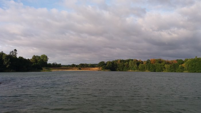 Molnigt landskap med en lugn sjö och trädbeklädda kustlinjer under ett tidigt morgonljus.