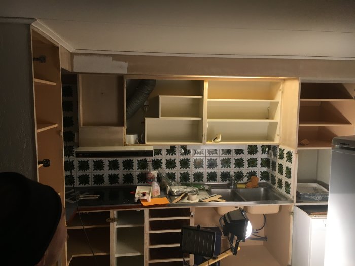 Kök under renovering med öppna, omålade träskåp och kakelväggar samt arbetsmaterial synliga.