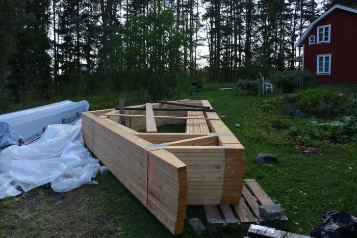 Nybyggda väggsektioner staplade på varandra i en trädgård med ett rött hus och skog i bakgrunden.