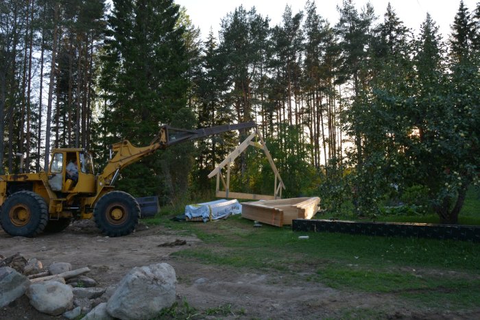 Byggplats med lastmaskin som lyfter takstol, stapel med råspont och omgivande träd.