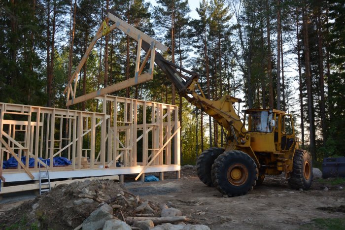 En lastmaskin lyfter en takstol på en halvfärdig trähusstomme i skogsmiljö.