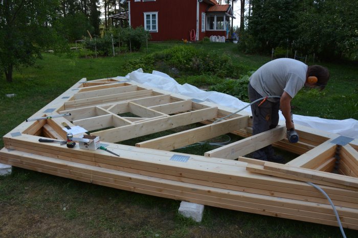 Man bygger en träkonstruktion utomhus med verktyg i förgrunden och ett rött hus i bakgrunden.