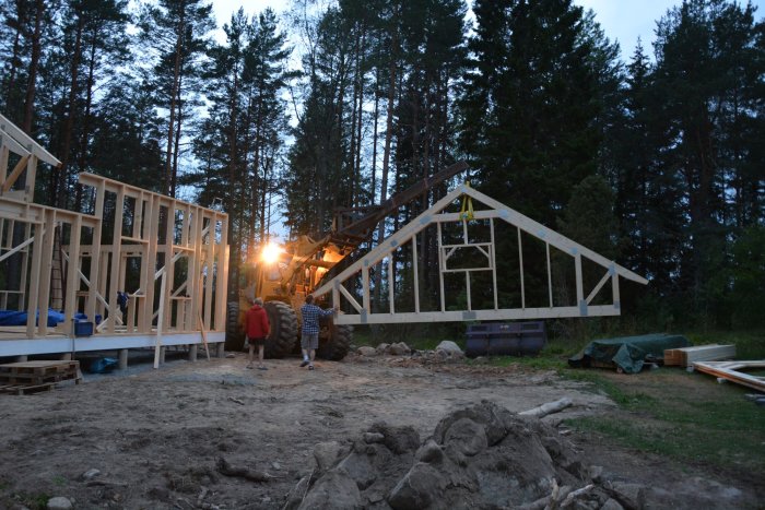 En hustomt vid skymningen med personer som arbetar på en nybyggd trästomme, takstolar som är delvis rester med hjälp av en lastmaskin.