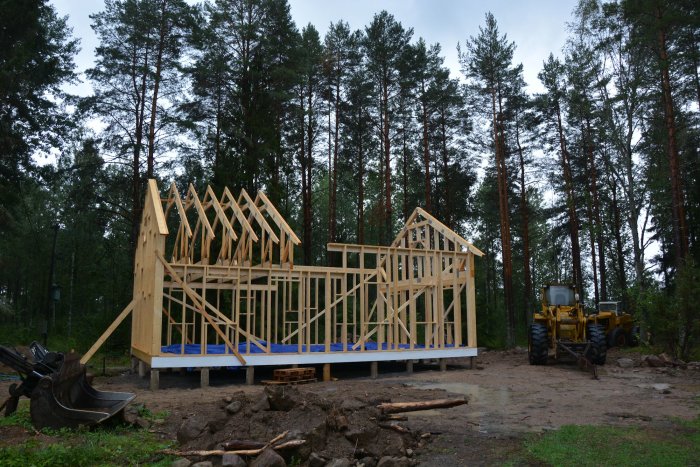 Under uppbyggnad av trähus med uppförda väggar och takstolar mot skogsbakgrund, med lastmaskin till höger.