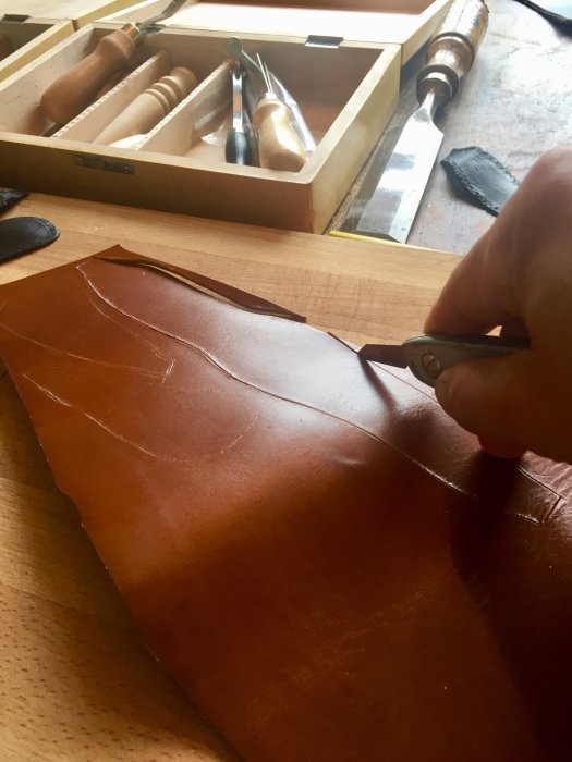 Hand skär till en ny läderhuva för växelspaksknopp på ett träbord med snideriverktyg i bakgrunden.