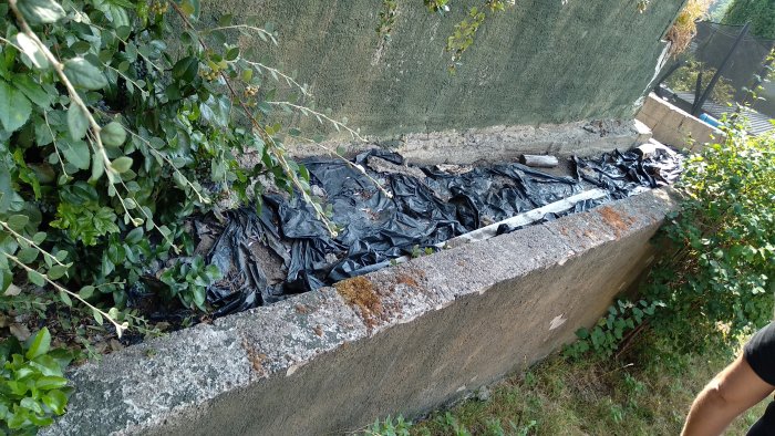 En gammal, sprucken stödmur i tre steg med utjämnad mark och plastduk, behöver renovering.