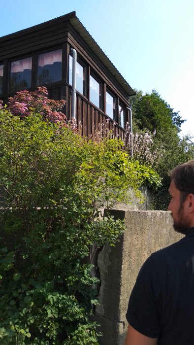 En villa med sutterängbyggnad, delvis skymd av grönska, med en gammal, delvis synlig, betongmur utanför.