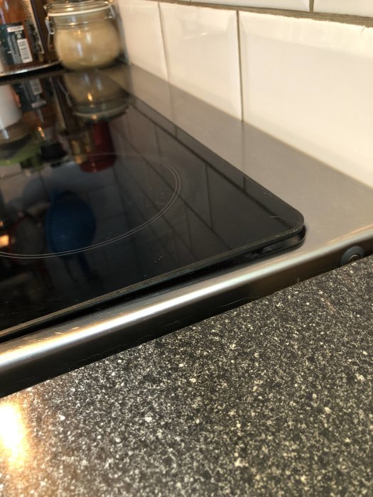 Häll i svart glaskeramik inbyggd i köksbänk av granit med kakelvägg i bakgrunden.
