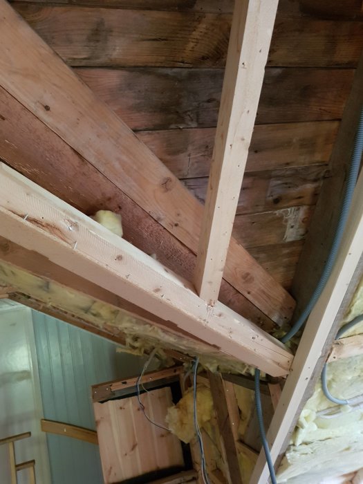 Interiör av vindsrum under renovering med exponerade takstolar, isolering och råspont som är mörkfärgad i hörnet.