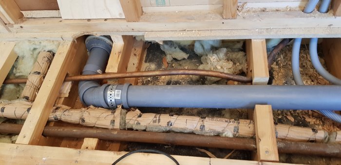 Upprivet trägolv som avslöjar rör och isolering, med ett grått avloppsrör stödd av en 45mm regel.