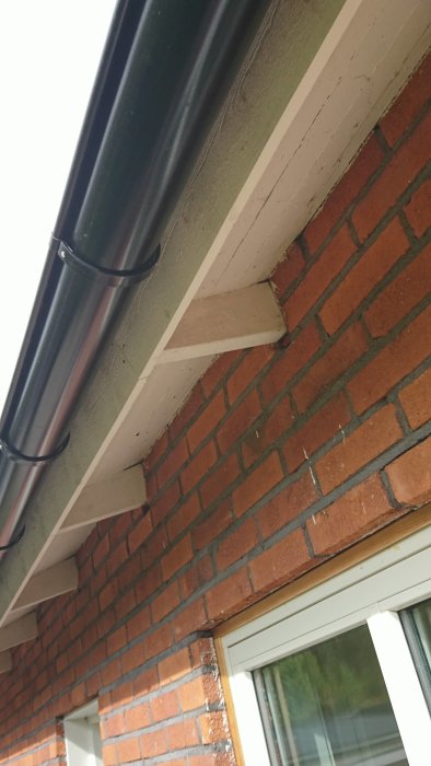 Stödkonsol för stuprör monterad på tegelfasad under takutsprånget av ett hus.