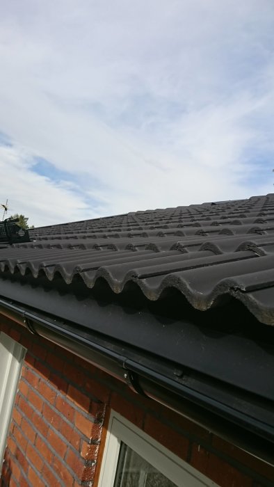Takpannor på ett hus med hängrännor, tegelfasad och ett fönster, under klart himmel.