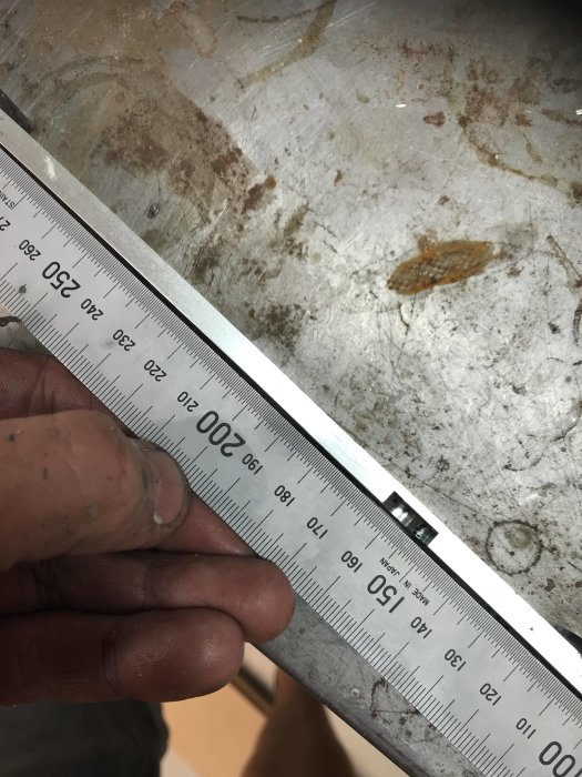 En hand håller en stålskena med tydliga mätmarkeringar mot en metallisk arbetsyta.
