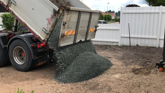 Lastbil tömmer grus på en uppfart under anläggning vid en vit staketbakgrund.