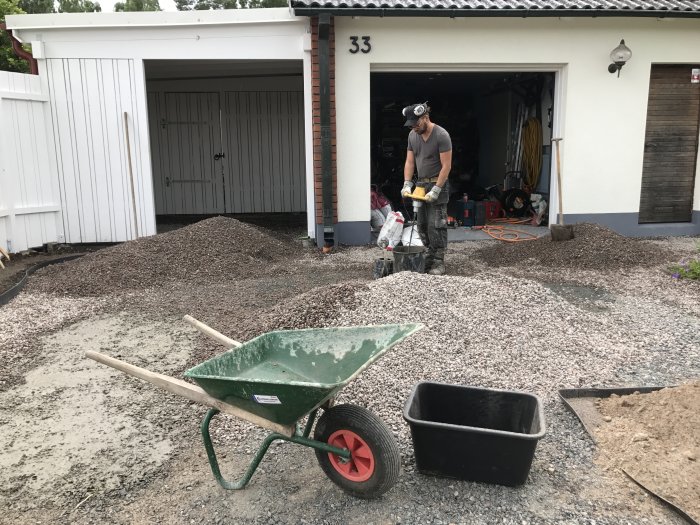 Person arbetar med att anlägga en ny uppfart med grus, sand, och byggmaterial framför ett garage.