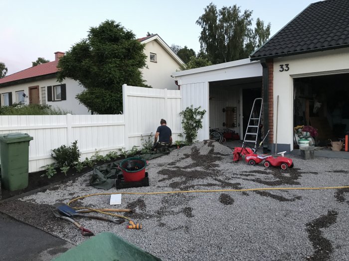 Person som arbetar på en uppfart med nylagda grus- och sandlager och verktyg utspridda, bredvid en vit staket och ett rött barnfordon.