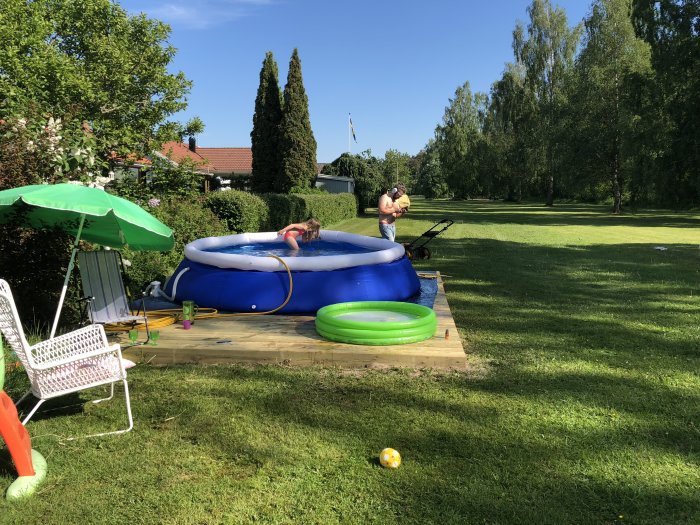 Trädgård med uppblåsbar pool på trädäck, gräsmatta, grillande person och grönska i bakgrunden.