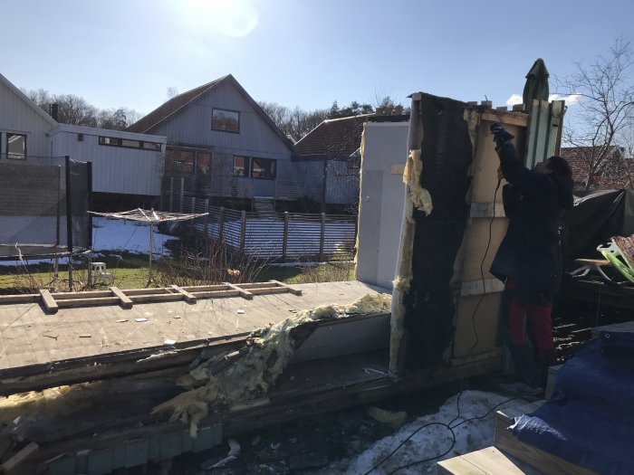 Rivning av gammalt förråd, person avlägsnar isolering, rester av byggmaterial på marken med hus i bakgrunden.