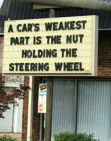 Skylt med texten "A car's weakest part is the nut holding the steering wheel" på en byggnads fasad.