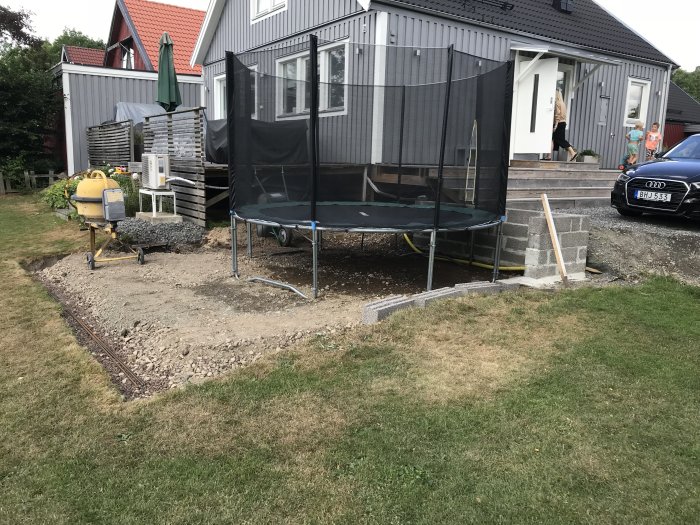 Förberedelse av mark för bygg med krossmaterial, stödmur och en trampolin framför ett hus.