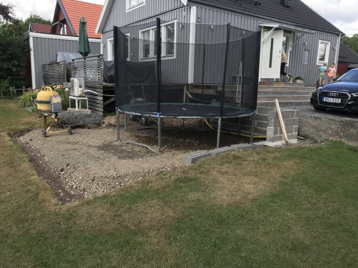 Förberedd mark för betongplatta vid hus med synlig grus, stödmur och trädgård.