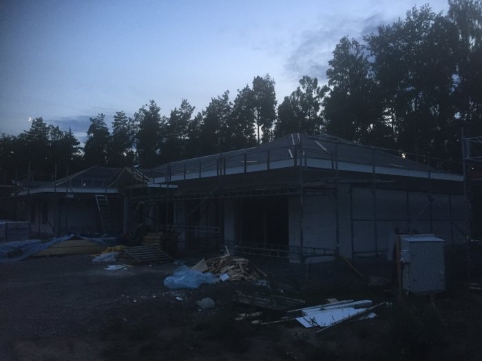 Kvällsbild av ett hus under konstruktion med delvis monterade takfotsbrädor och byggmaterial på marken.