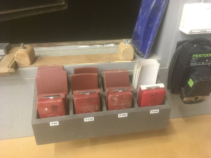Grå hållare med små slipklossar i olika grovlekar märkta P80, P120, P180, P240 på en arbetsbänk.