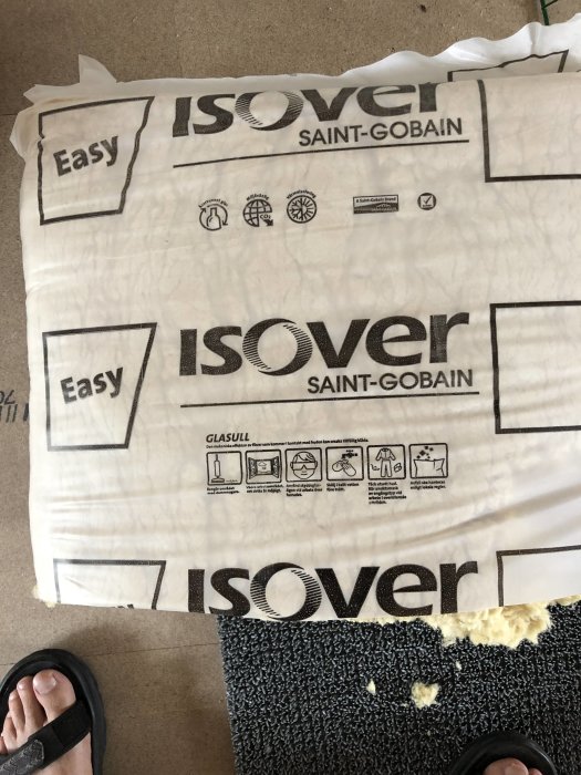 Förpackning av lösullsisolering märkt 'ISOVER SAINT-GOBAIN Easy' med instruktioner, delvis rullad på ett golv vid en persons fötter.
