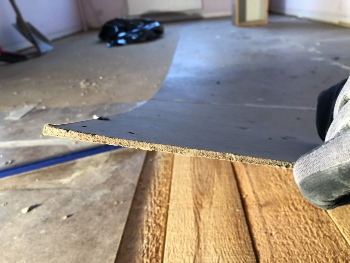 Närbild på en hand som lyfter en masonitskiva och avslöjar underliggande granplankor på ett golv under renovering.