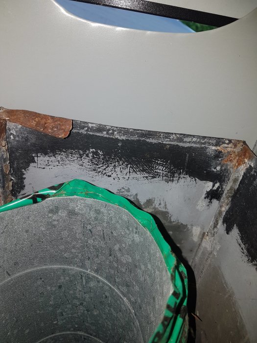 En öppen byggnadshuv med synliga skruvhål och obruten tätning, närbild på grov och rostig kant.