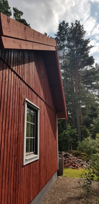 Ett rött trähus med nymålat gavelsprång, omgivet av en skogsträdgård och en hög med gamla tegelstenar.