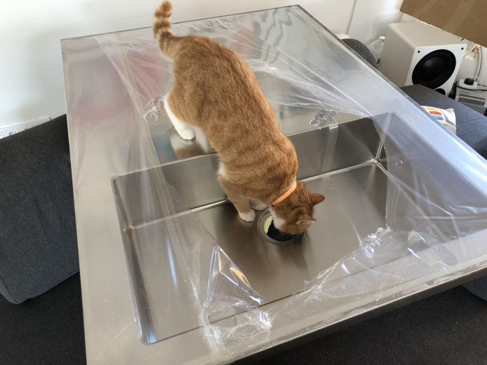 Katt inspekterar en ny 90 cm bred diskbänk inslagen i plast som ligger på golvet.