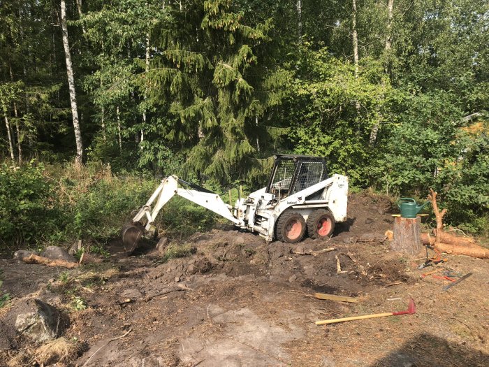 En grävmaskin schaktar marken vid en skogsbryn för förberedelse av attefallshusbygge.