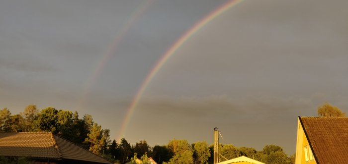 Dubbel regnbåge över ett bostadsområde vid solnedgång med hus och träd.