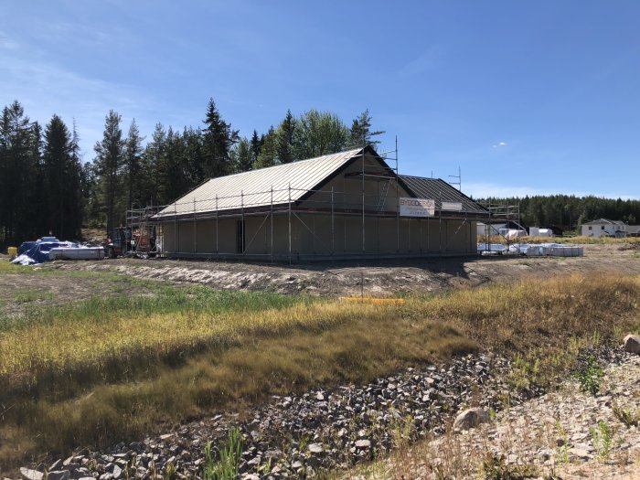 Hus under konstruktion med stommen uppe och taket på plats, omgivet av en byggarbetsplats och skog.