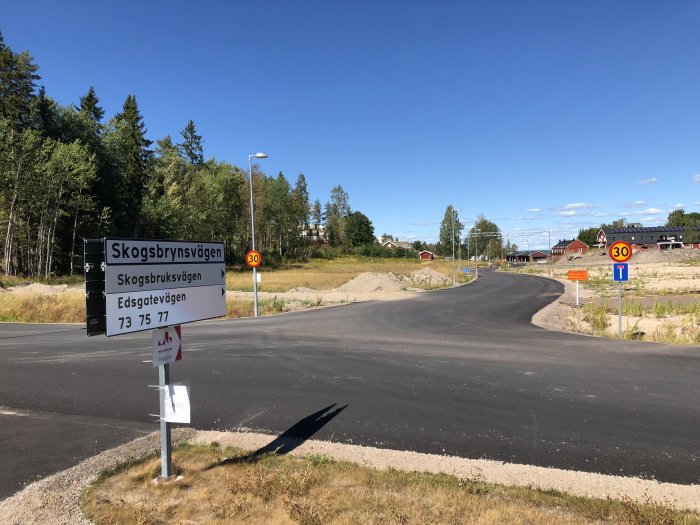 Nybyggt bostadsområde med gatuskyltar och några hus i bakgrunden under en klarblå himmel.