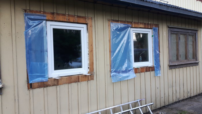Renovering av husfasad med nyligen utbytta fönster skyddade av plast och en äldre fönsterram.
