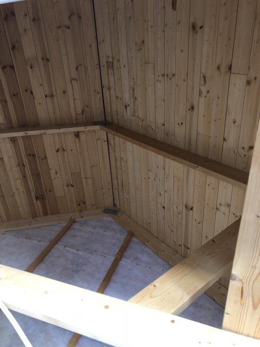 Inre hörn av ett trähus under konstruktion med synliga sparrar och isolering.