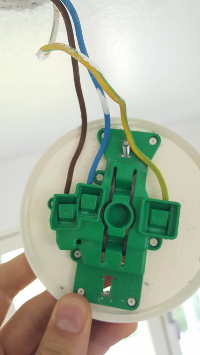 En hand som håller en elektrisk kopplingsdosa till taklampa med synliga ledningar och kopplingar.