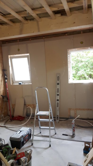Renoveringsarbete pågår med nytt ytterväggsgips, öppet tak och fönster samt verktyg och stege på golvet.
